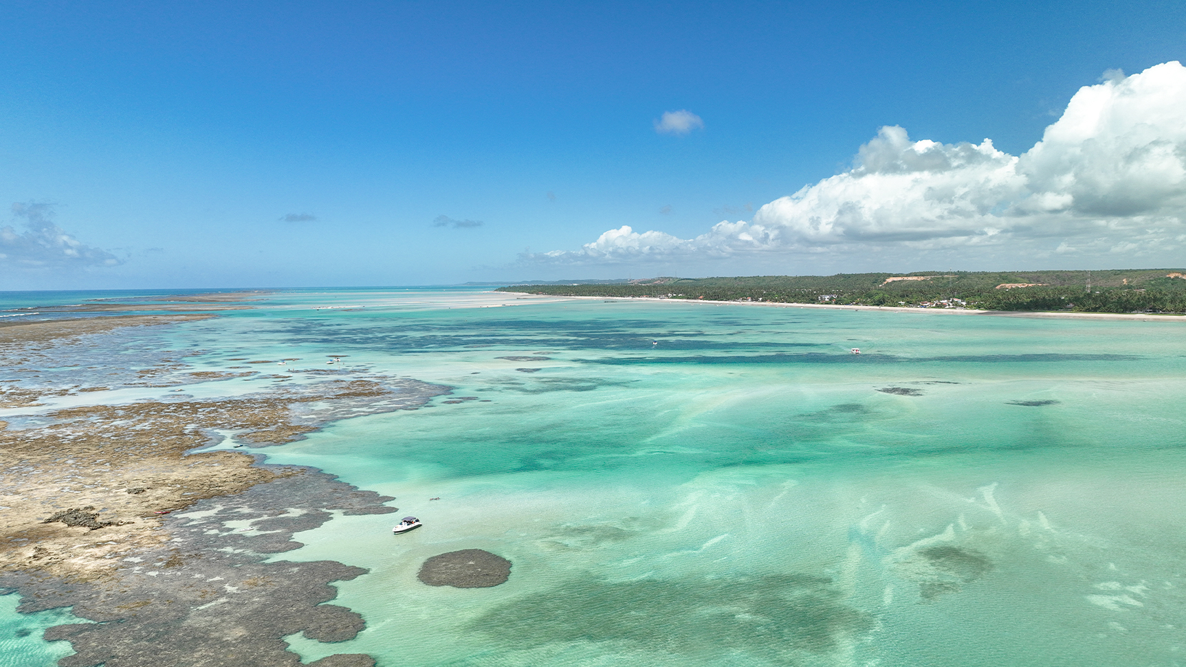 Vista aérea da praia Ponta de Mangue, com recifes de corais e águas cristalinas das piscinas naturais e há um barco ancorado na prainha.