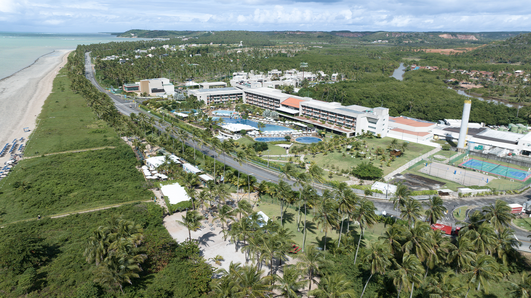 Vista aérea do Japaratinga Lounge Resort à beira-mar, com piscinas amplas e quadras, cercado por vegetação e coqueiros. E no canto esquerdo um é possível ver a praia de Japaratinga.