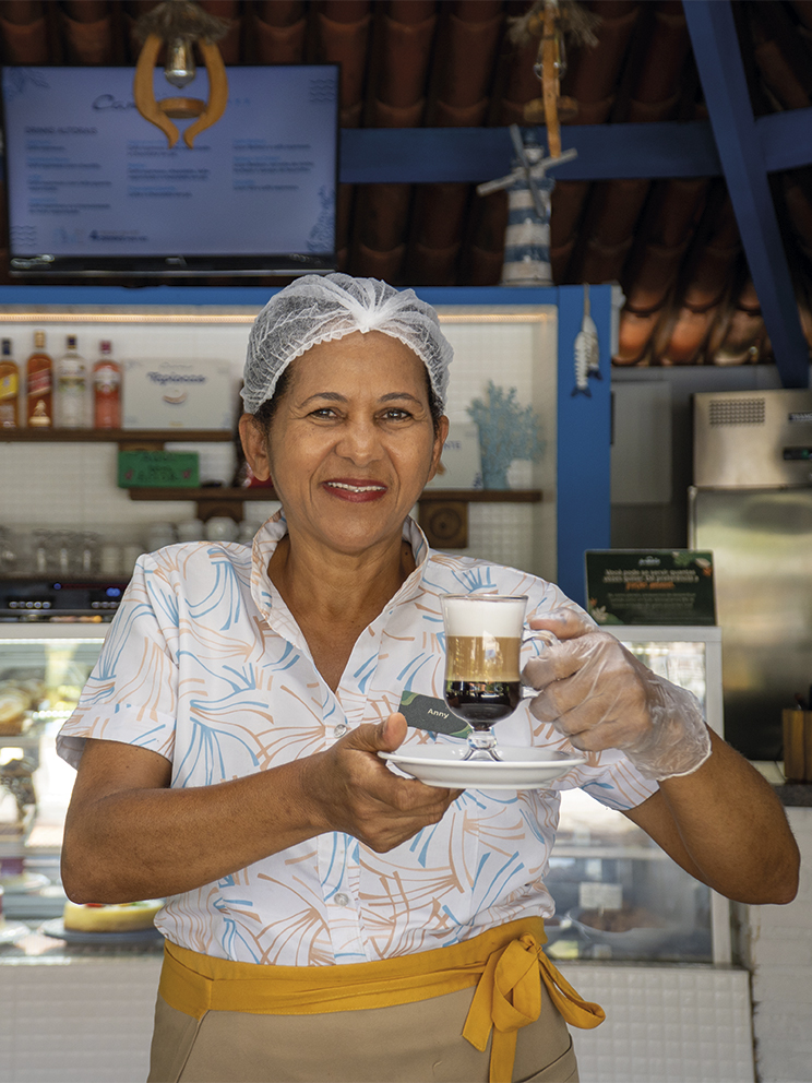Café e doces na experiência All Inclusive Pessoa colaboradora do Salinas Maragogi servindo café especial no Café Canoas com vitrine de sobremesas ao fundo.