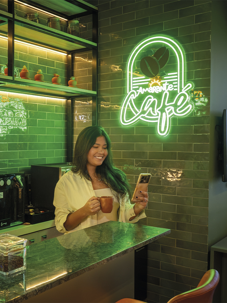 Amarante Café o conforto pra dar energia Mulher sorridente segura uma xícara de café e olha pro celular em um balcão moderno. O ambiente possui azulejos verdes, prateleiras de madeira com plantas e um letreiro neon escrito Amarante Café.