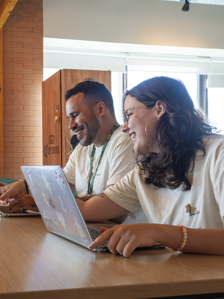 Construímos juntos na Amarante Dois colaboradores da Amarante sorriem enquanto trabalham juntos em notebooks em uma mesa de madeira. Ao fundo, parede de tijolinhos com o logotipo A estilizado e luminárias pendentes modernas.