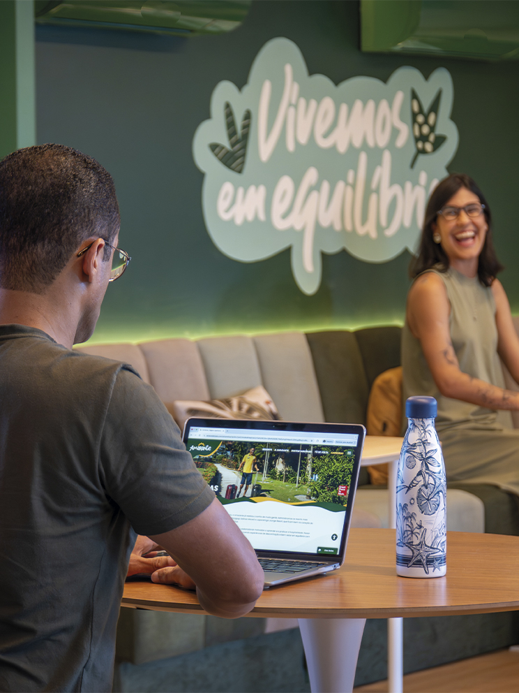 Bem-estar Amarante trabalhando com equilíbrio Ambiente de coworking com parede verde onde se lê Vivemos em equilíbrio. Um colaborador em primeiro plano e uma colaboradora ao fundo sorriem enquanto trabalham em um espaço acolhedor.