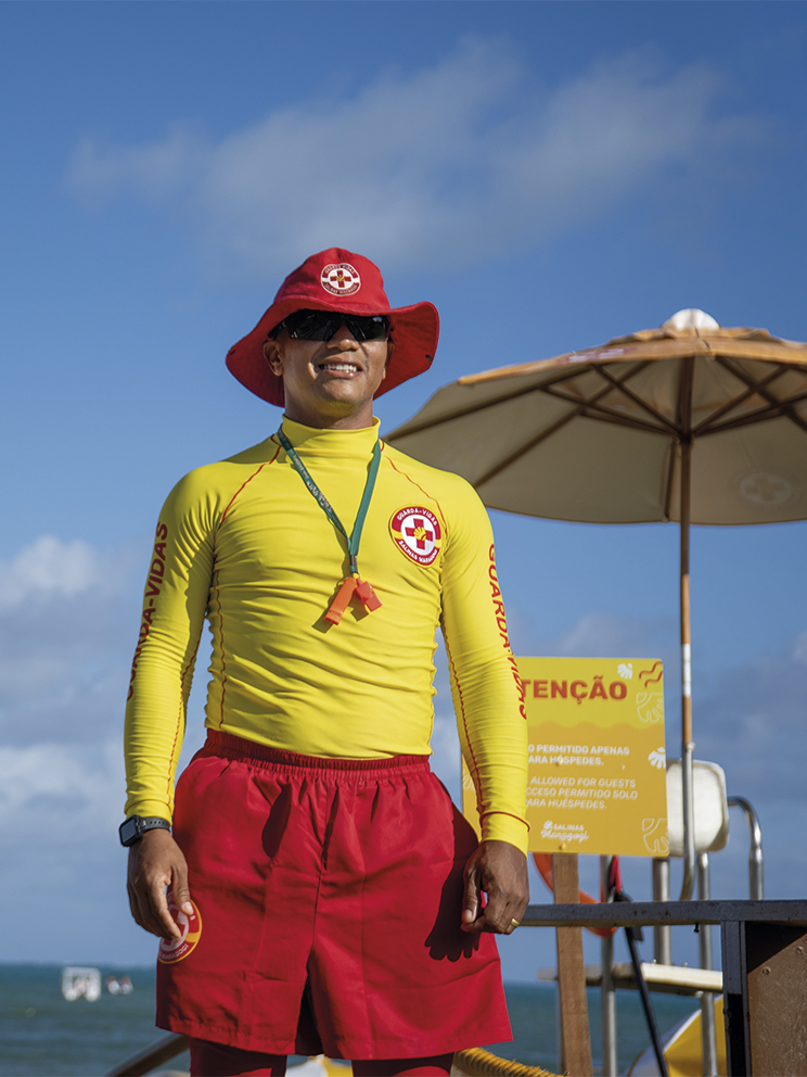 Segurança em primeiro lugar no Salinas Maragogi Salva-vidas do Salinas Maragogi sorri sob o sol, vestindo uniforme amarelo e vermelho com chapéu protetor. Ao fundo, o mar azul, céu com nuvens e guarda-sóis amarelos de praia.