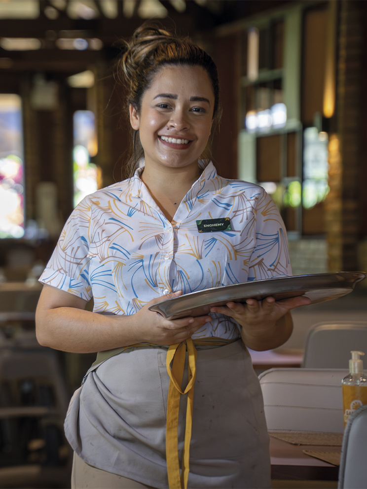 O sorriso que acolhe no Salinas Maragogi Colaboradora sorridente segura uma bandeja em um ambiente do restaurante em Salinas Maragogi. Ela veste uniforme estampado e o fundo mostra um salão com iluminação suave e detalhes em madeira.