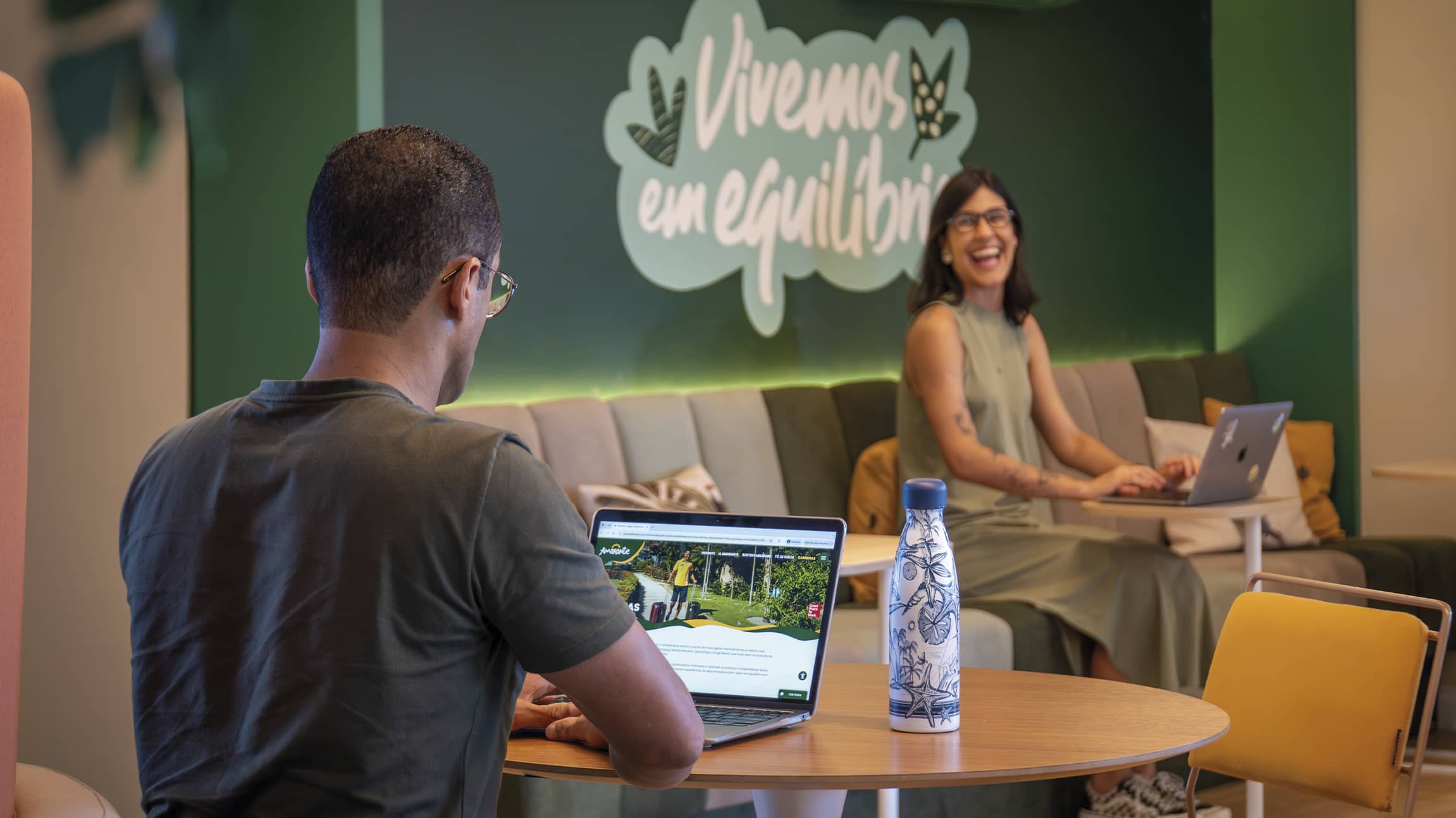 Bem-estar Amarante trabalhando com equilíbrio Ambiente de coworking com parede verde onde se lê Vivemos em equilíbrio. Um colaborador em primeiro plano e uma colaboradora ao fundo sorriem enquanto trabalham em um espaço acolhedor.