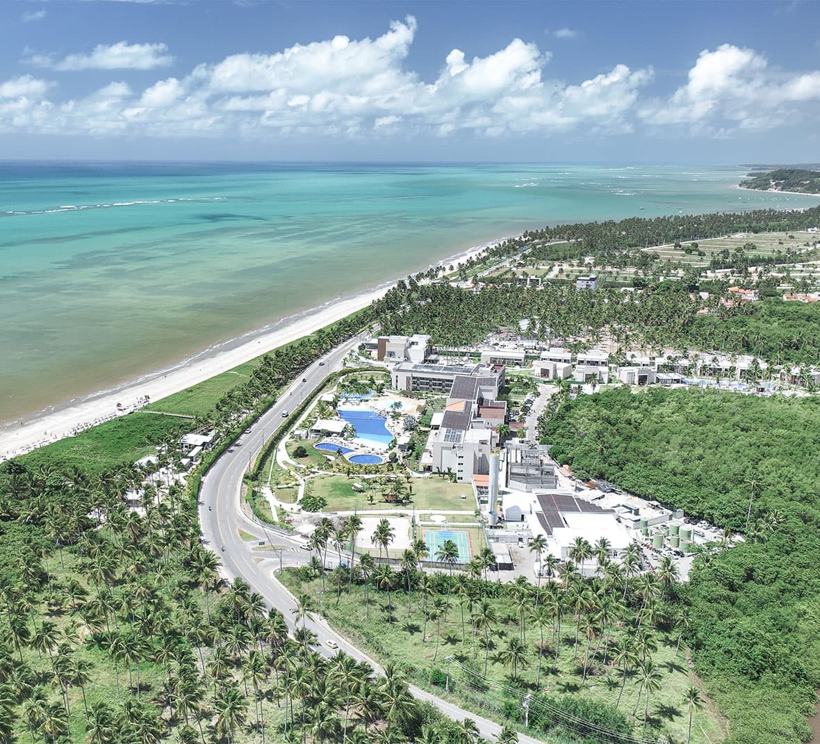 Vista aérea de um resort Japaratinga Lounge Resort, piscinas e quadras, situado entre uma extensa praia de areia e um rio de mangue. A área é cercada por um vários coqueiros e o mar é de um azul-turquesa intenso.