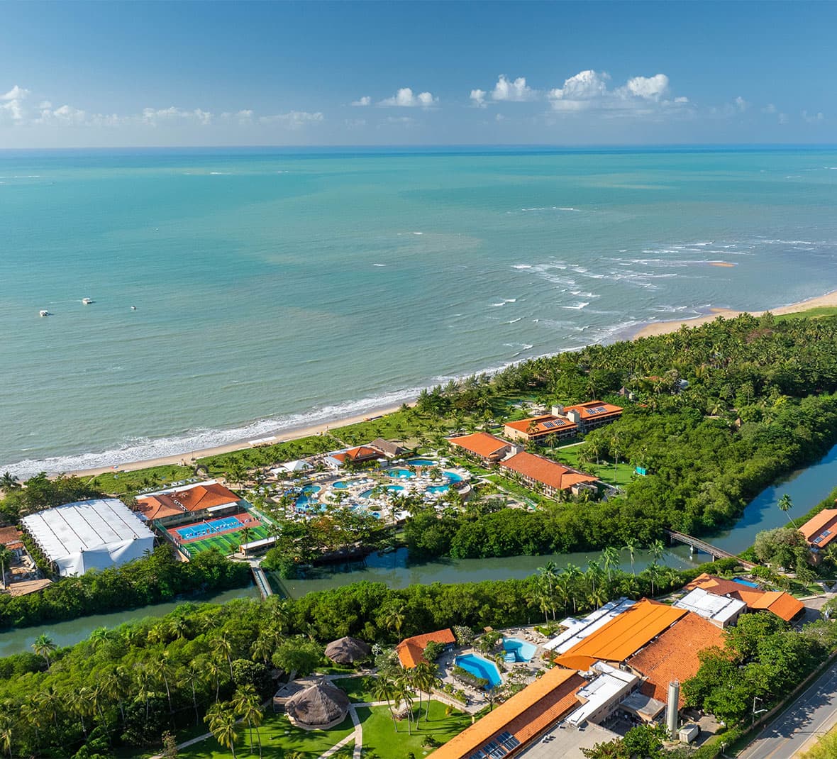 Vista de ângulo elevado do complexo do Salinas Maragogi, mostrando uma área de lazer infantil, quadras, piscinas maiores, estrutura do resort, com o rio cortando o resort com uma ponte ligando os dois lados, e o mar azul com a praia ao fundo, tudo cercado por coqueiros.