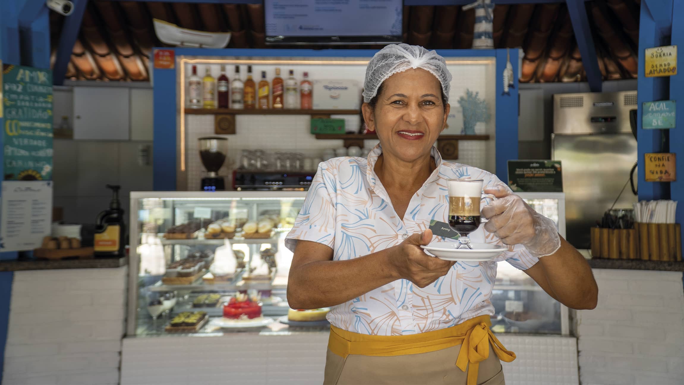Café e doces na experiência All Inclusive Pessoa colaboradora do Salinas Maragogi servindo café especial no Café Canoas com vitrine de sobremesas ao fundo.