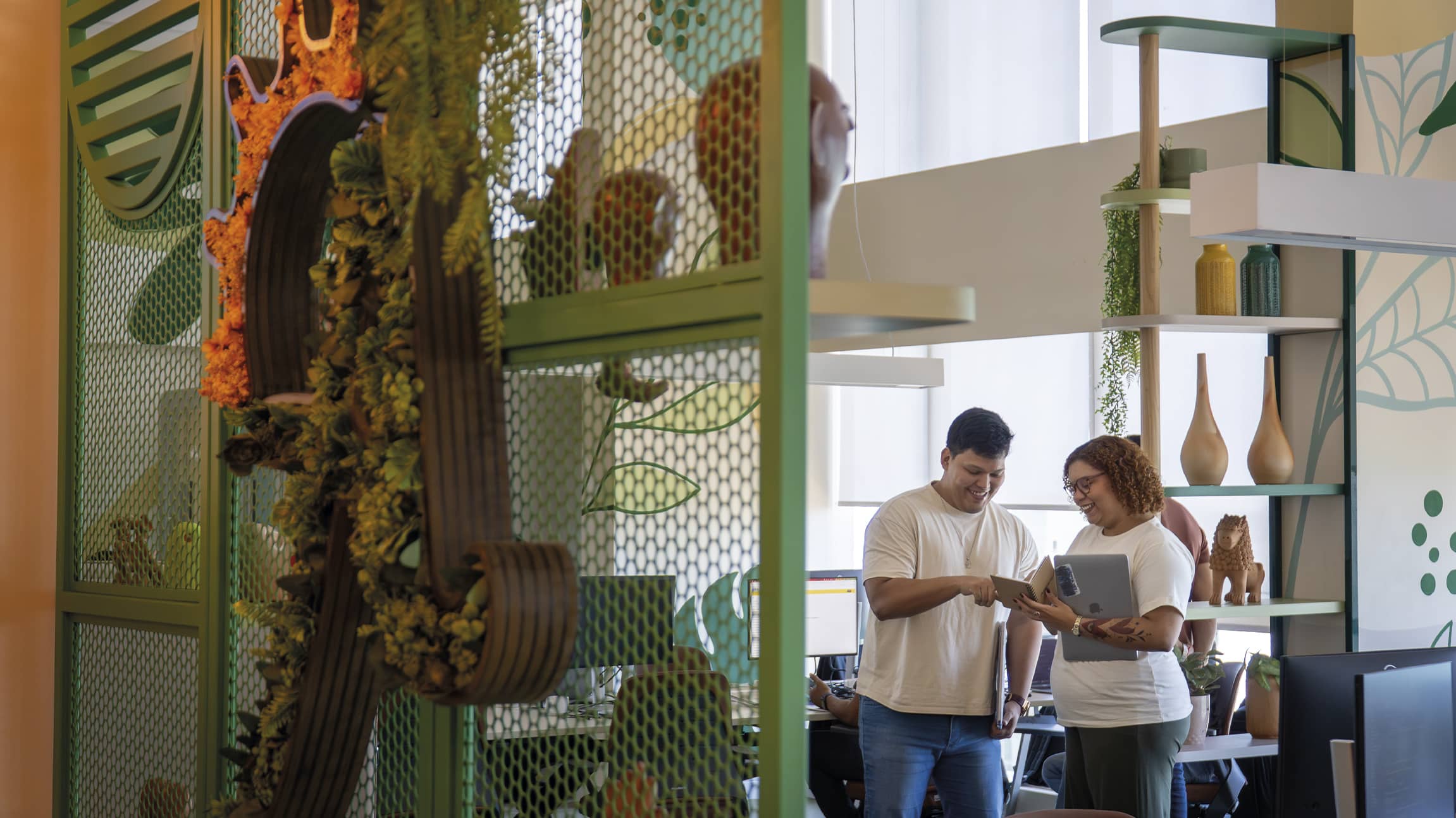 Cultura Amarante em ambiente de trabalho Colaboradores da Amarante sorriem enquanto conversam em um escritório moderno. Em primeiro plano, uma divisória verde com plantas e elementos orgânicos, ao fundo, um ambiente iluminado com decoração colorida mais verde.