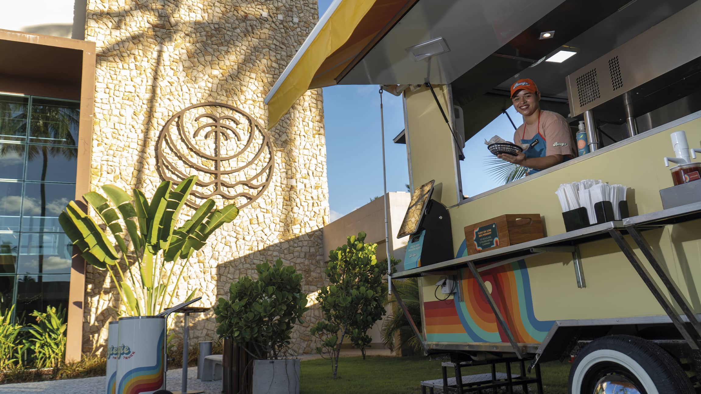 Boogie Truck no Japaratinga Lounge Resort Colaboradora sorridente atende em um food truck colorido no Japaratinga Lounge Resort, entregando um lanche. Ao fundo, uma parede de pedras claras exibe o logotipo do Japaratinga Lounge Resort sob a luz do sol e sombras de palmeiras.