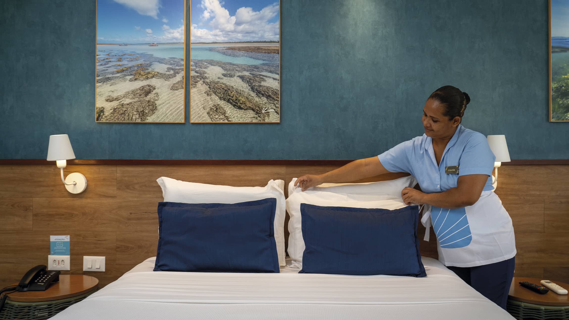 Cuidado e conforto em cada detalhe Colaboradora da governança do Salinas Maceió, usando uniforme azul e branco, arruma cuidadosamente os travesseiros de uma cama de casal em um quarto decorado com tons de azul e madeira.