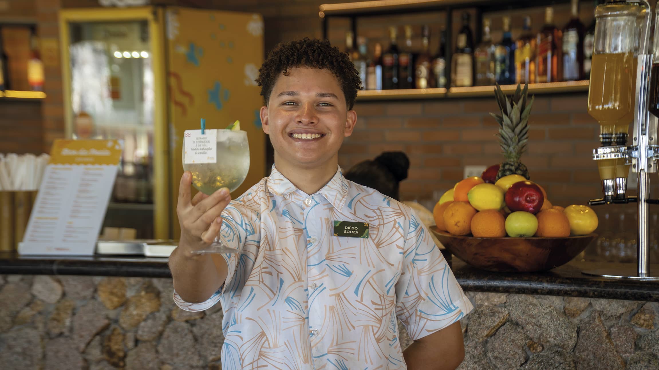 Drinks e alegria, o jeito Salinas Maragogi O colaborador do Salinas Maragogi, sorri segurando uma taça de drink refrescante em frente a um bar de pedras. Ele veste uma camisa estampada e ao fundo há uma fruteira tropical colorida.