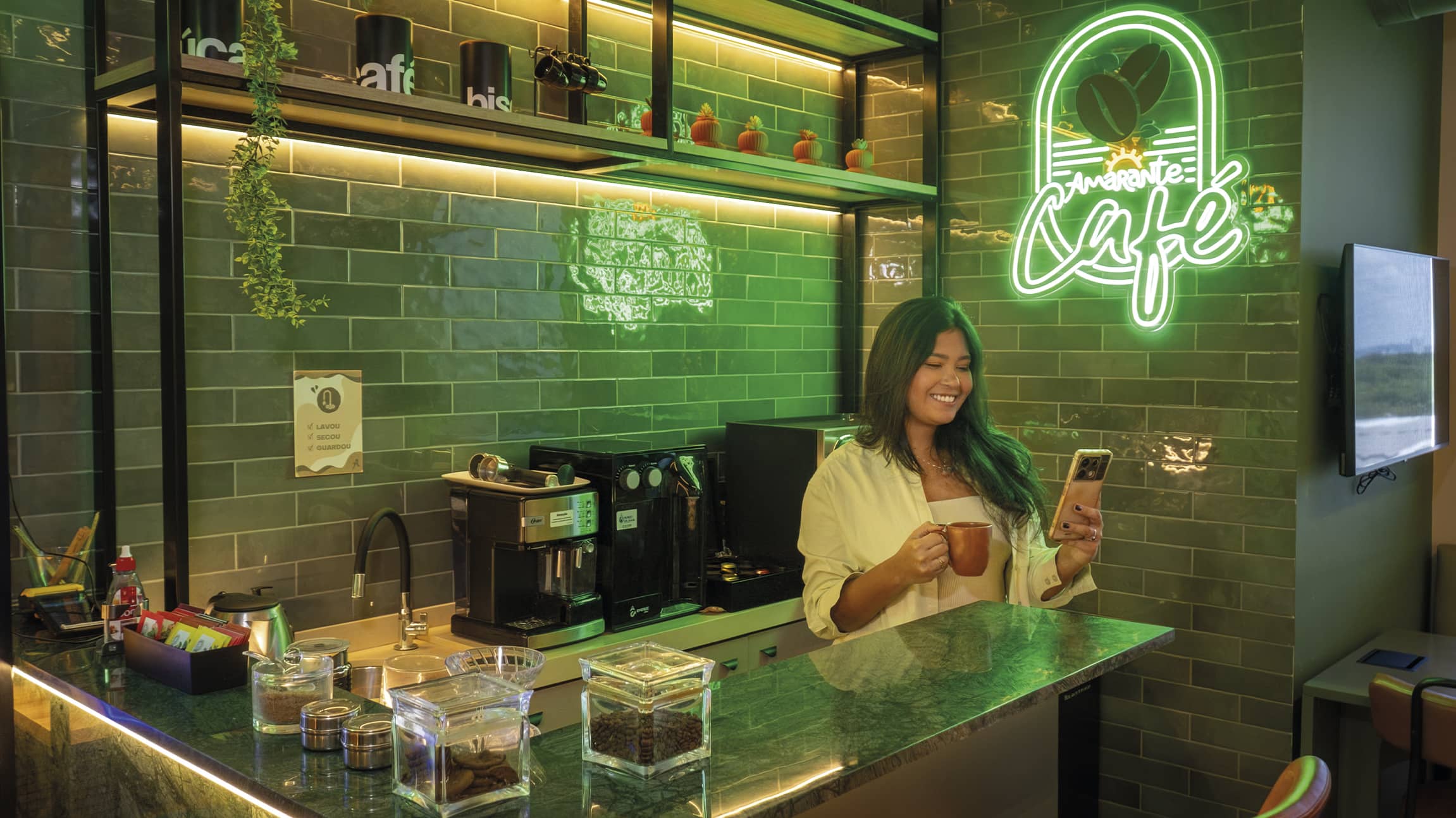 Amarante Café o conforto pra dar energia Mulher sorridente segura uma xícara de café e olha pro celular em um balcão moderno. O ambiente possui azulejos verdes, prateleiras de madeira com plantas e um letreiro neon escrito Amarante Café.