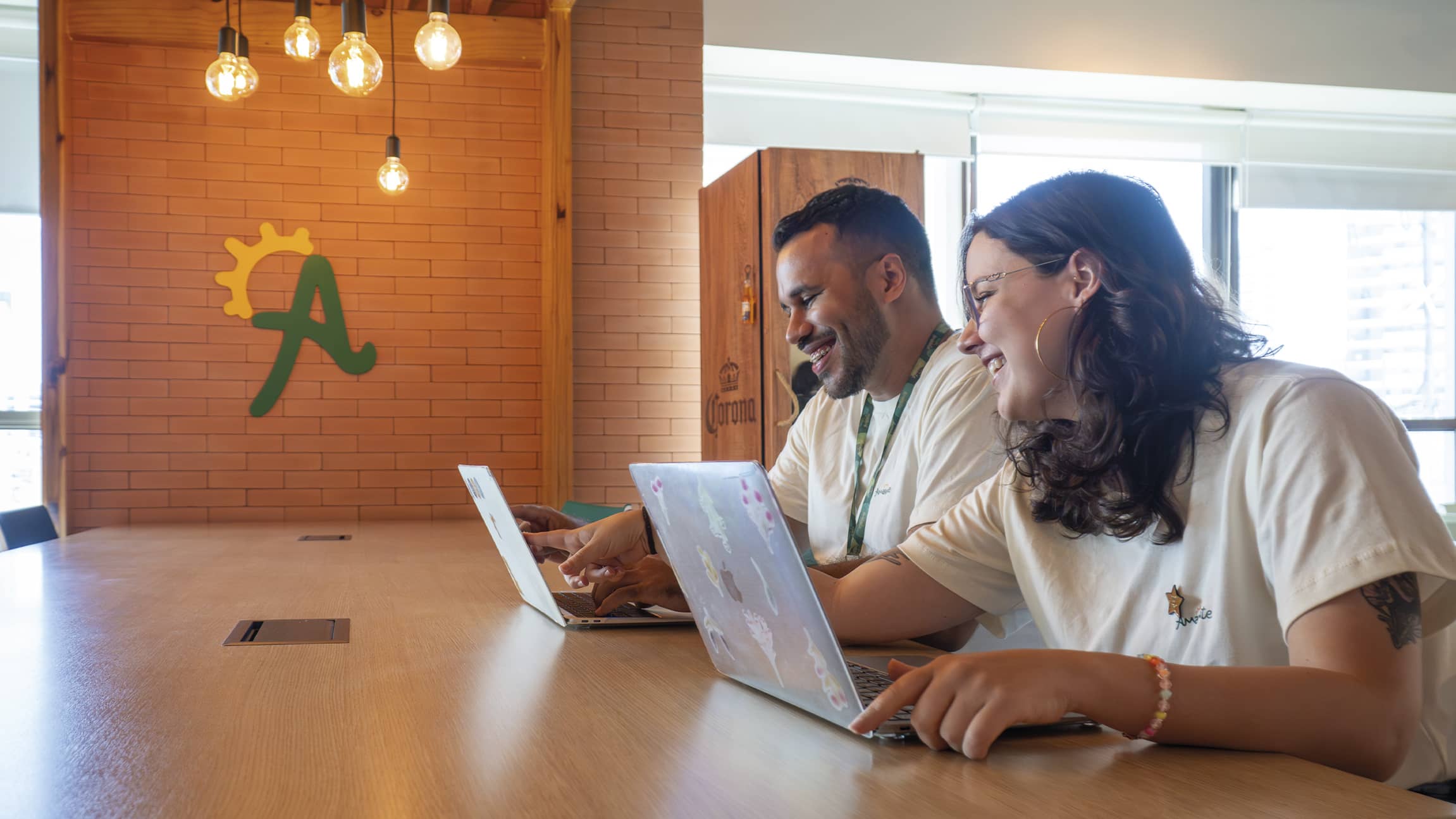 Construímos juntos na Amarante Dois colaboradores da Amarante sorriem enquanto trabalham juntos em notebooks em uma mesa de madeira. Ao fundo, parede de tijolinhos com o logotipo A estilizado e luminárias pendentes modernas.