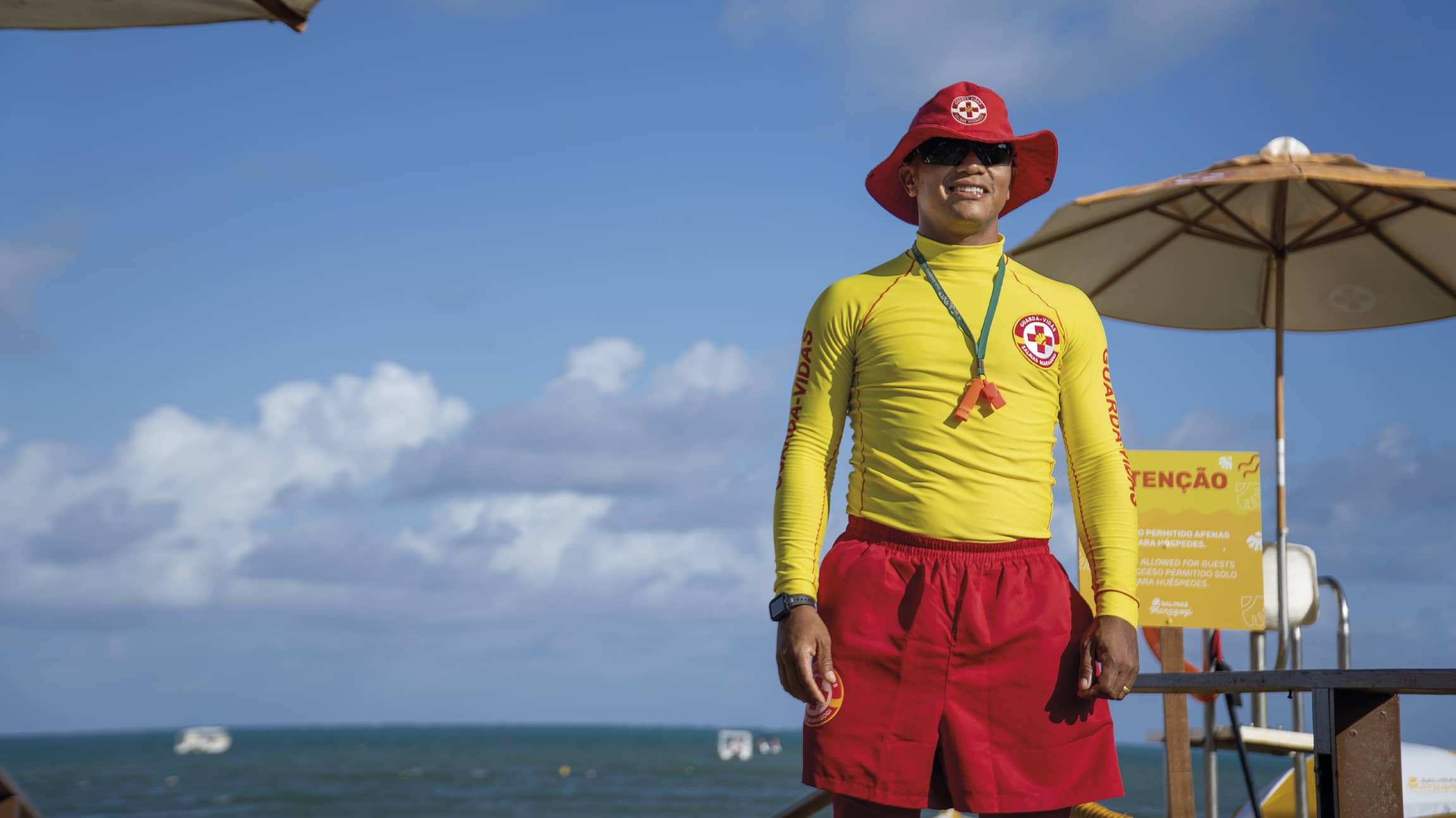 Segurança em primeiro lugar no Salinas Maragogi Salva-vidas do Salinas Maragogi sorri sob o sol, vestindo uniforme amarelo e vermelho com chapéu protetor. Ao fundo, o mar azul, céu com nuvens e guarda-sóis amarelos de praia.
