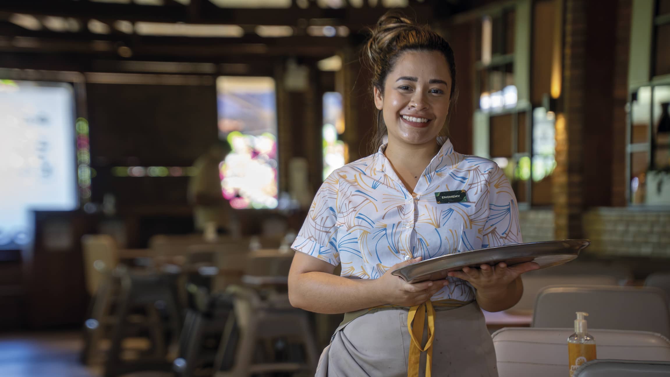O sorriso que acolhe no Salinas Maragogi Colaboradora sorridente segura uma bandeja em um ambiente do restaurante em Salinas Maragogi. Ela veste uniforme estampado e o fundo mostra um salão com iluminação suave e detalhes em madeira.