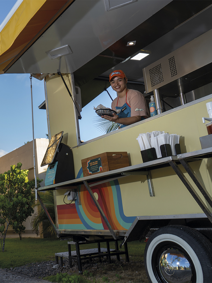 Boogie Truck no Japaratinga Lounge Resort Colaboradora sorridente atende em um food truck colorido no Japaratinga Lounge Resort, entregando um lanche. Ao fundo, uma parede de pedras claras exibe o logotipo do Japaratinga Lounge Resort sob a luz do sol e sombras de palmeiras.