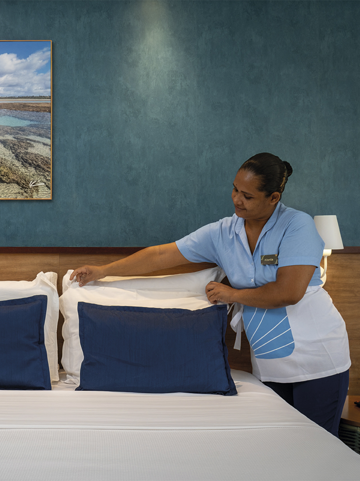 Cuidado e conforto em cada detalhe Colaboradora da governança do Salinas Maceió, usando uniforme azul e branco, arruma cuidadosamente os travesseiros de uma cama de casal em um quarto decorado com tons de azul e madeira.
