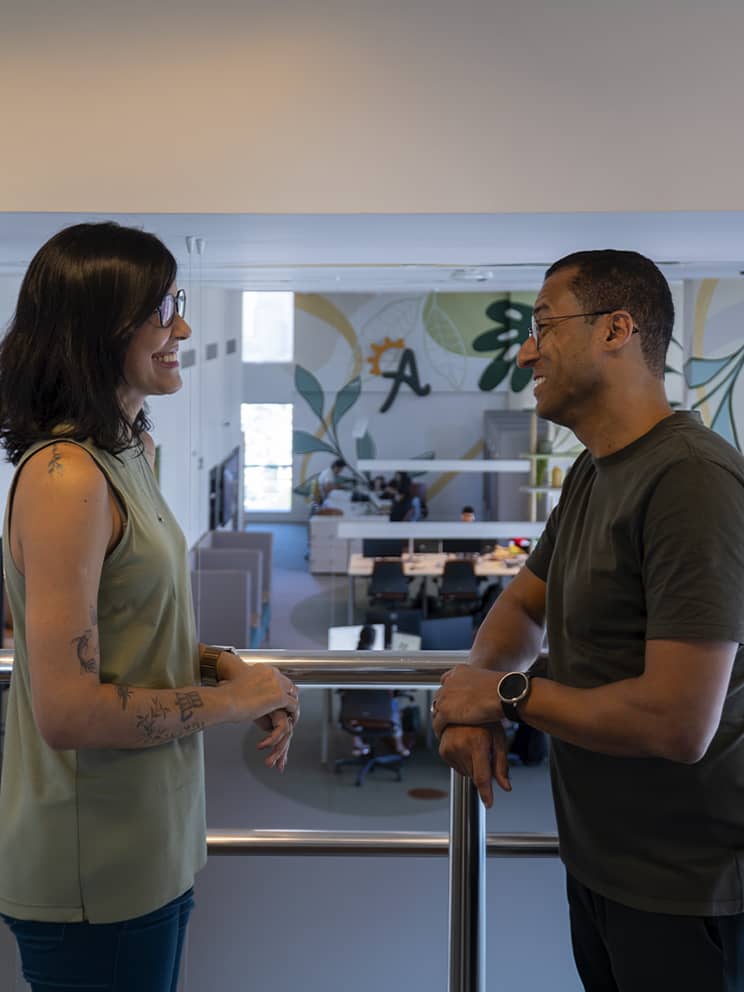 Ambiente de trabalho colaborativo na Amarante Pessoas colaboradoras da Amarante interagindo no escritório de Recife. Decoração com plantas e um ambiente leve e acolhedor. Uma grande que separa a recepção do escritório tem a logo da Amarante.