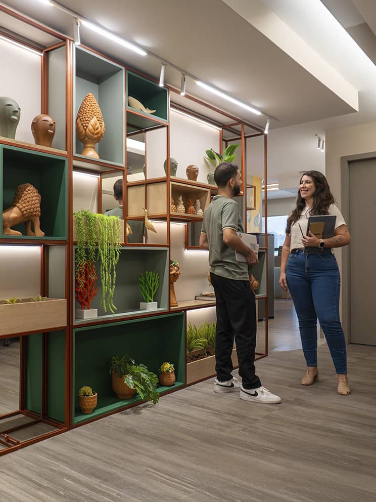 Espaço interno do escritório corporativo da Amarante Corredor do escritório corporativo da Amarante com duas colaboradoras andando e conversando, ao lado direito um letreiro da Amarante e uma baia de trabalho com funcionários e computadores, e ao lado esquerdo uma prateleira de decoração.