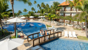 Vista aérea das piscinas e blocos de apartamentos do Salinas Maceió cercados por coqueirais sob céu azul. A família está na piscina infantil na área de lazer.