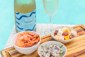 A garrafa de Moscatel do Salinas Maceió com taça ao lado. Petiscos como camarão, ceviche e queijos variados à beira da piscina.