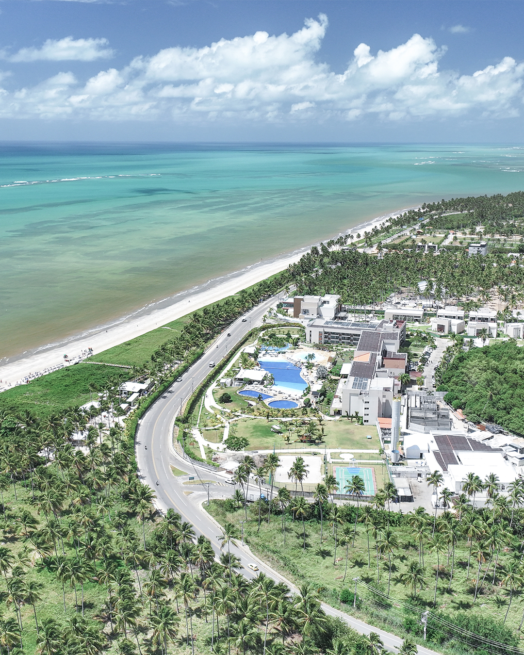 Vista aérea de um resort Japaratinga Lounge Resort, piscinas e quadras, situado entre uma extensa praia de areia e um rio de mangue. A área é cercada por um vários coqueiros e o mar é de um azul-turquesa intenso.
