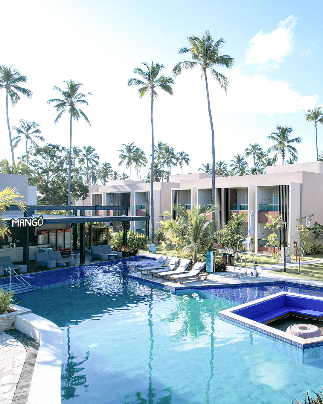 Vista da piscina do Japaratinga Lounge Resort, com coqueiros altos e os quartos Gardens ao fundo. A piscina tem uma área submersa com assentos azuis, o Mangô Bar é visível à esquerda e o céu está com poucas nuvens.