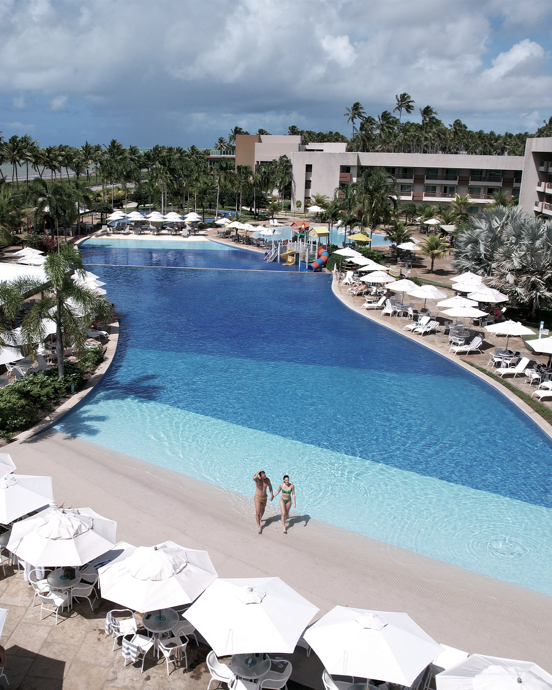 Piscina principal do Japaratinga Lounge Resort. Um casal de mãos dadas caminha na parte rasa. A piscina é rodeada por inúmeros guarda-sóis e espreguiçadeiras brancas e coqueiros. Há o bar Verano no canto e a estrutura do Japaratinga Lounge Resort.