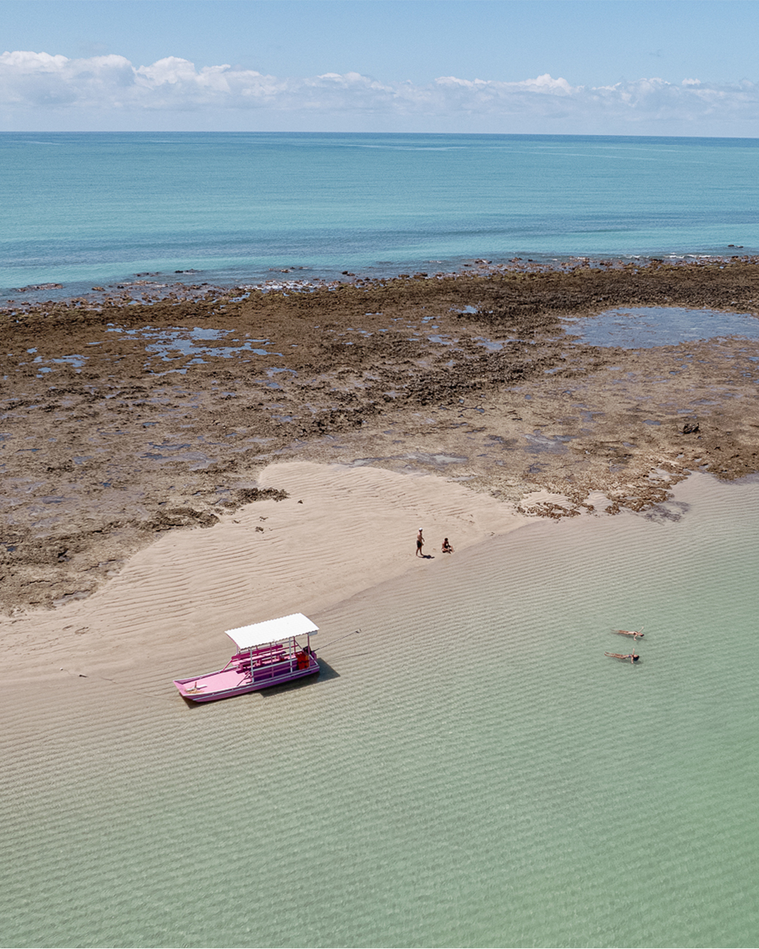 Um barco rosa com duas pessoas na areia onde ele está ancorado em águas rasas e cristalinas. Há uma longa faixa de recifes de corais, separando o mar azul cristalino de um mais turquesa ao fundo. e o céu esta com poucas nuvens.