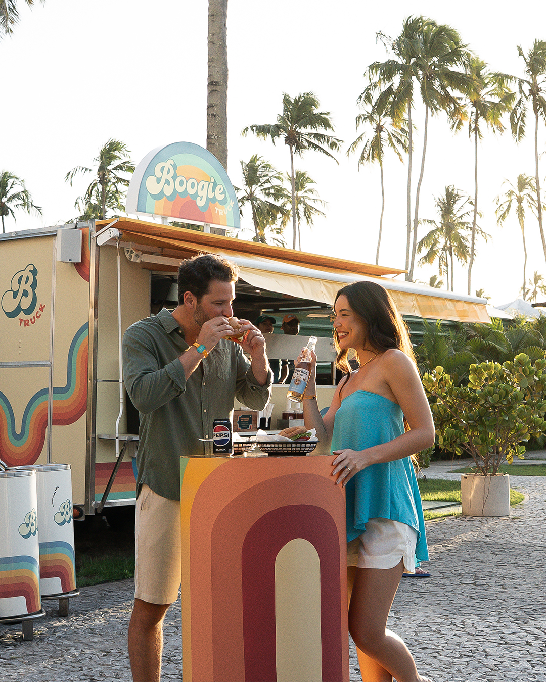 Casal jovem sorrindo em frente a um food truck Boogie. O homem está comendo um hambúrguer e a mulher está com uma Corona na mão. O ambiente é ao ar livre, cercado por coqueiros sob a luz dourada do fim de tarde.