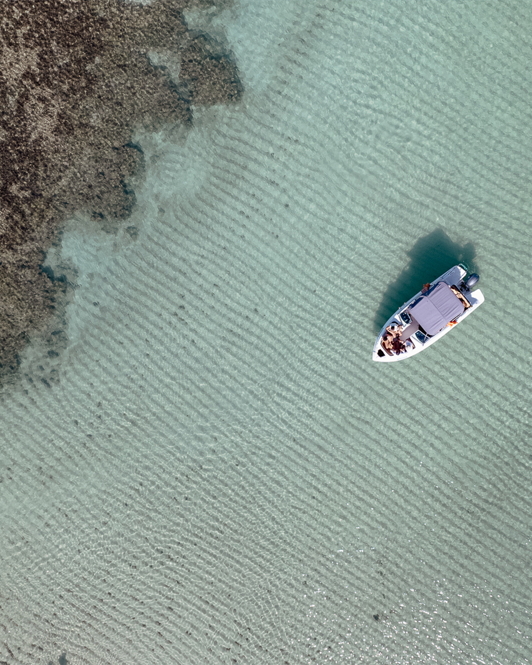Vista aérea de uma lancha branca, ancorada em águas verde-claras e cristalinas. Ao redor, há inúmeras formações de recifes de corais em tons de marrom escuro, caracterizando as piscinas naturais de Japaratinga. Um homem que anda pelas piscinas é visível no canto inferior esquerdo.