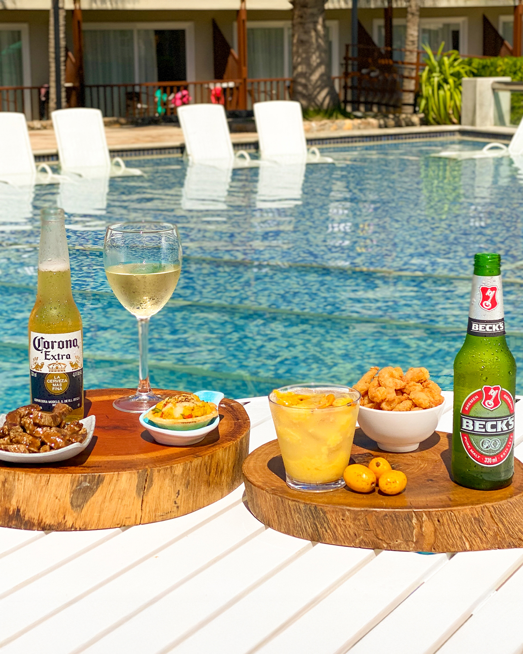 Mesa branca com duas tábuas de madeira servindo petiscos, uma garrafa de Corona, uma de Beck's, um vinho branco e um drink em copo. A mesa está posicionada à beira da piscina azul do resort Salinas Maceió.