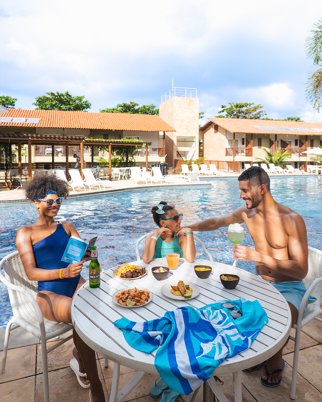Família sorrindo, sentados em uma mesa branca à beira da piscina grande. Eles estão comendo petiscos e bebendo drinks. Ao fundo, a piscina, espreguiçadeiras e a arquitetura do resort Salinas Maceió sob o sol.