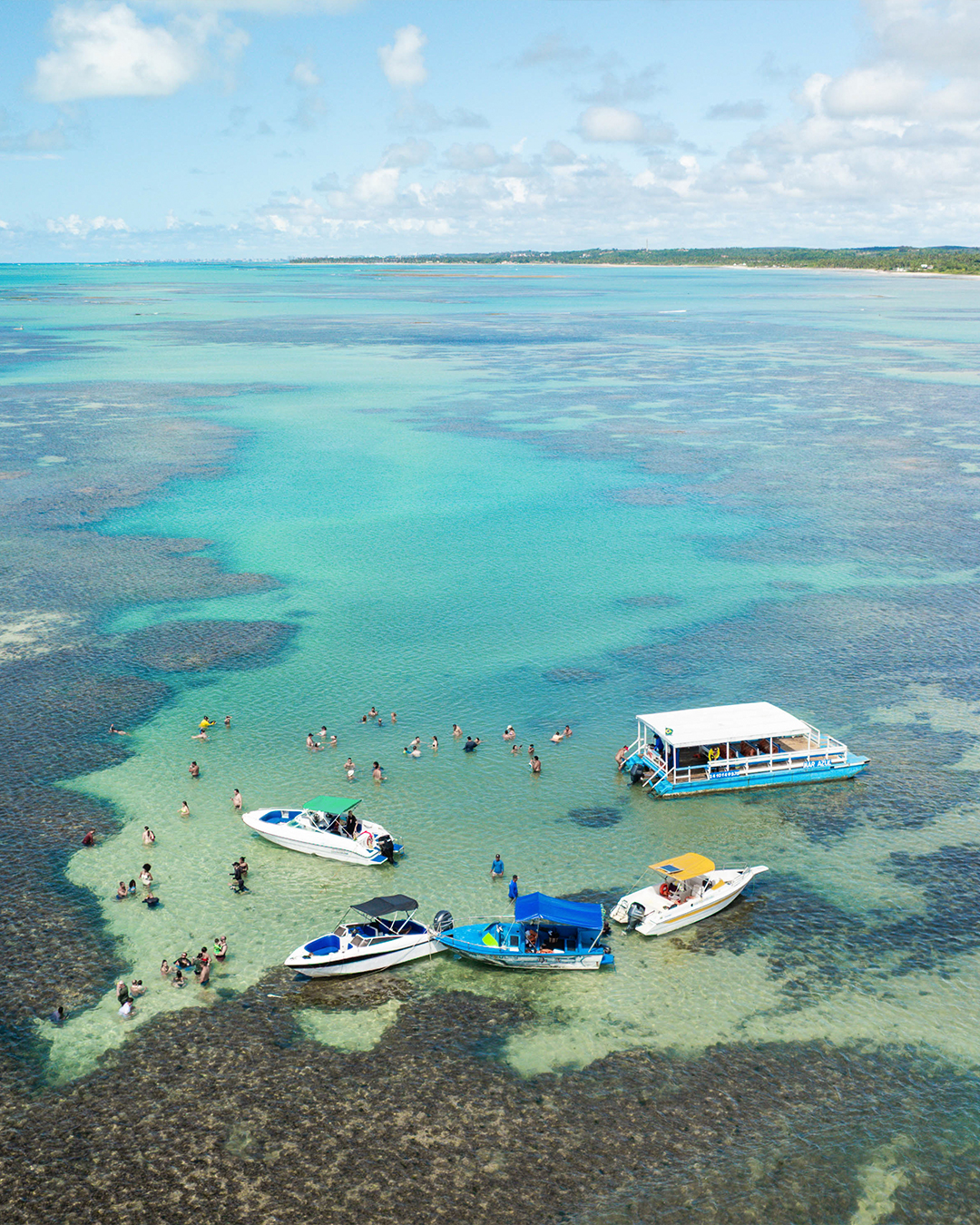Vista aérea de piscinas naturais com grandes formações de recifes de corais. Cinco embarcações e muitos turistas são visíveis aproveitando as águas cristalinas de tom azul-turquesa. O horizonte mostra a costa litorânea e céu com poucas nuvens.