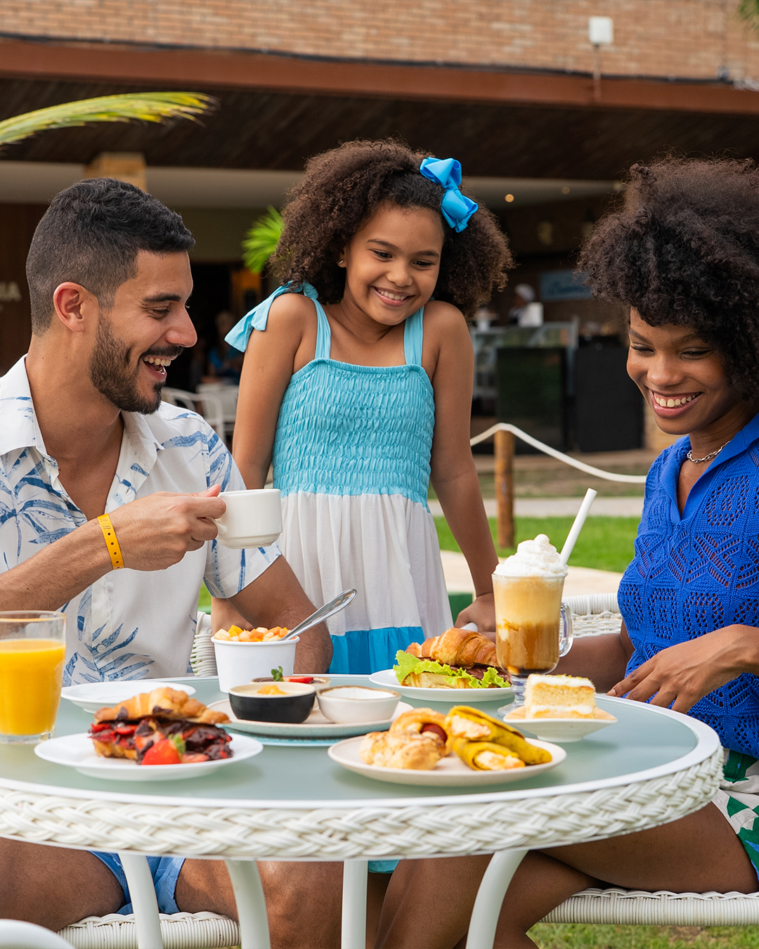 Família sorrindo, sentada em mesas brancas no gramado do resort Salinas Maceió, desfrutando de um lanche com sanduíches, salgados, doces e bebidas. O pai segura uma caneca de café, a mãe uma bebida gelada e a filha observa.