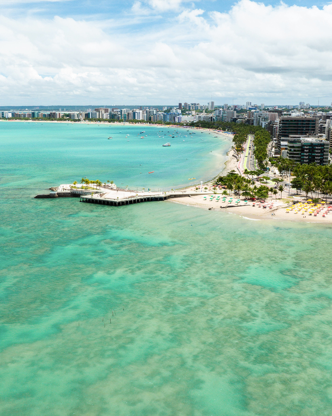 Vista aérea panorâmica da orla urbana de Maceió. Uma longa praia de areia clara com guarda-sóis coloridos e pessoas, um píer de concreto e o mar azul-turquesa com recifes visíveis. Edifícios altos se estendem pela costa.