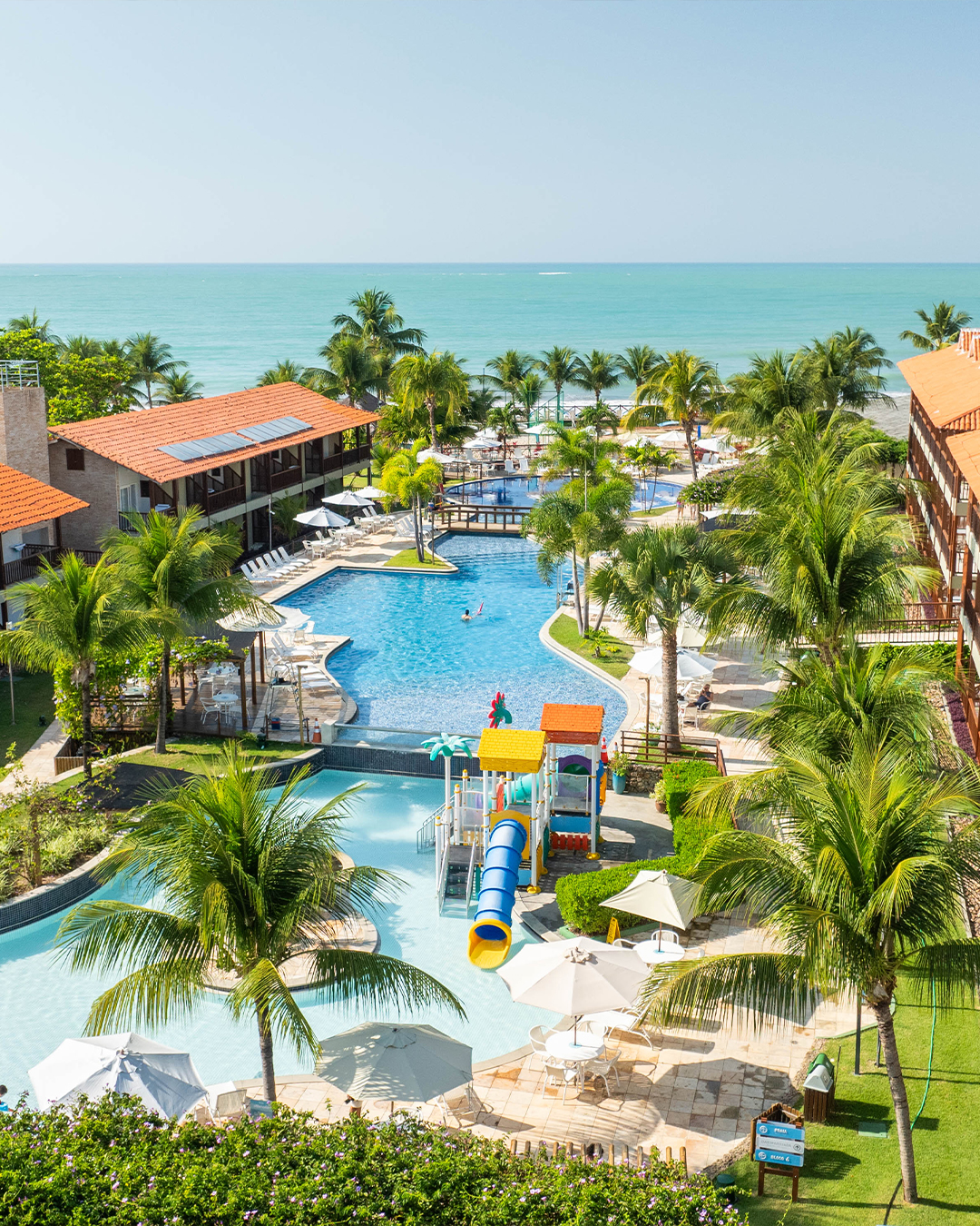 Vista aérea de um resort Salinas Maceió pé na areia com prédios de telhados laranjas. A área de lazer inclui piscinas com decks, coqueiros e um parque aquático infantil com escorregadores coloridos. O mar está visível ao fundo.