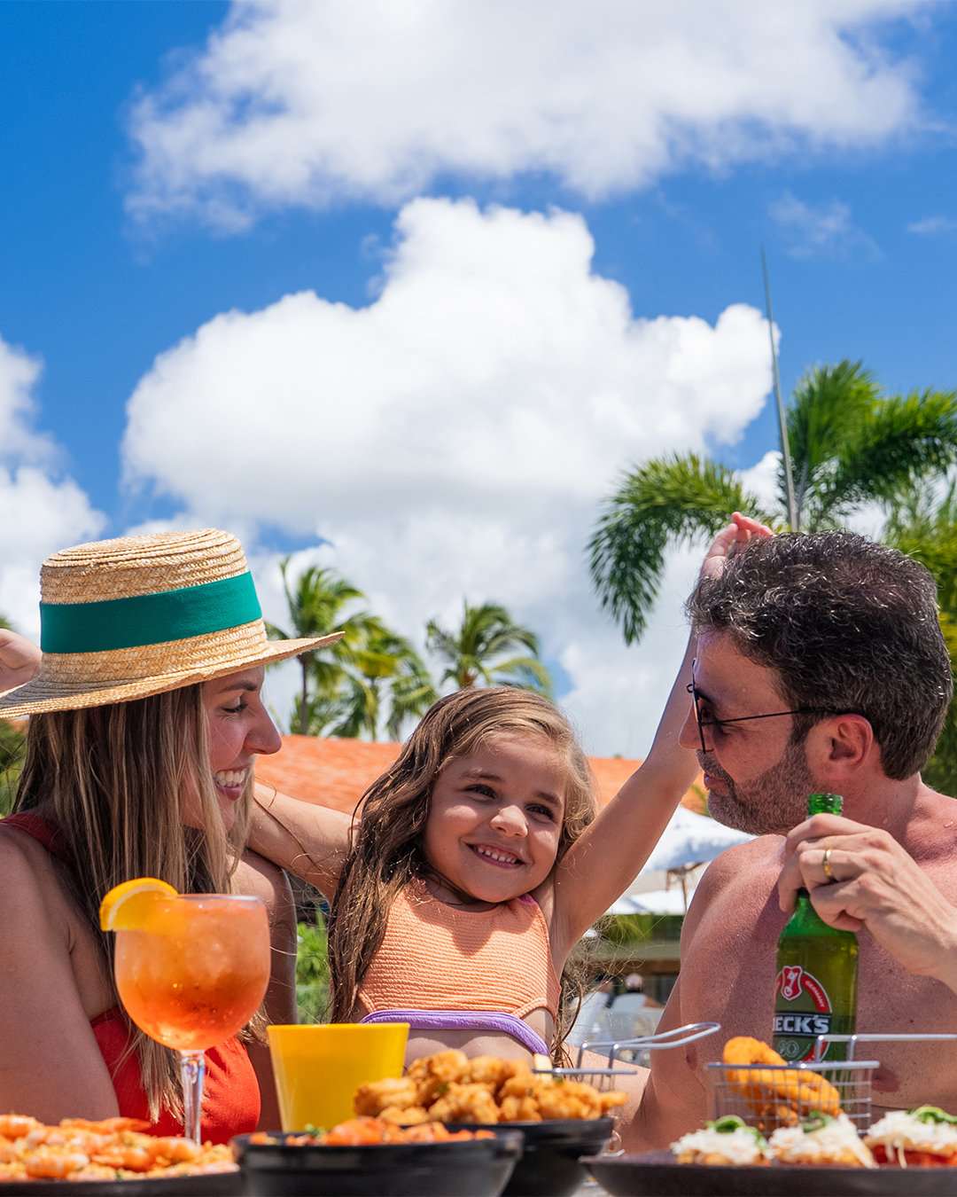 Família sorridente com filha abraçada com pai e mãe em uma mesa com diversos pratos de comida e bebidas, ao lado da área da piscina. Ao fundo, algumas espreguiçadeiras, guarda-sóis, coqueiros e a estrutura do Salinas Maragogi.