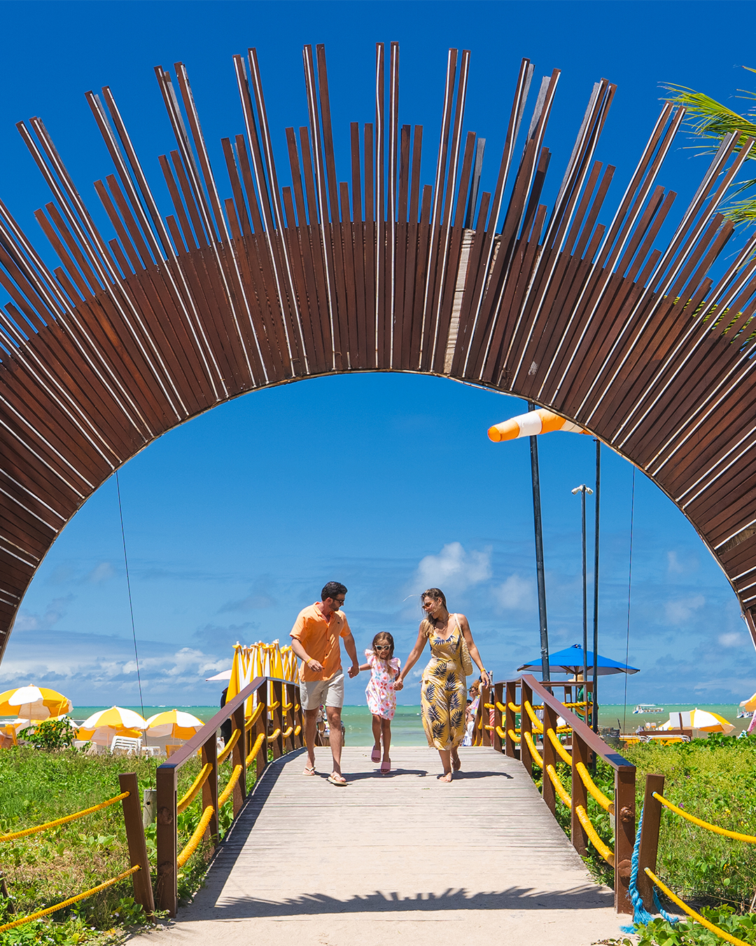 Família com pai, mãe e filha pequena de mãos dadas, caminhando sorridentes em direção à câmera, passando por dentro de um grande arco de madeira que serve de portal pra a praia do resort, com coqueiros e o céu azul ao fundo.