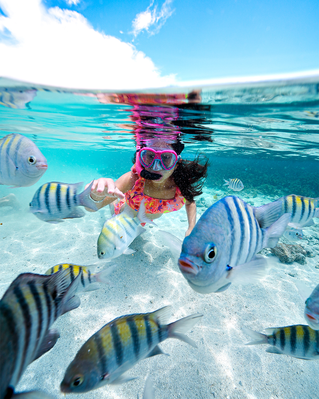 Foto meio submersa de uma menina usando máscara de snorkel, rodeada por peixes listrados em águas cristalinas. O fundo de areia branca é visível, e a superfície da água reflete o céu azul com nuvens.
