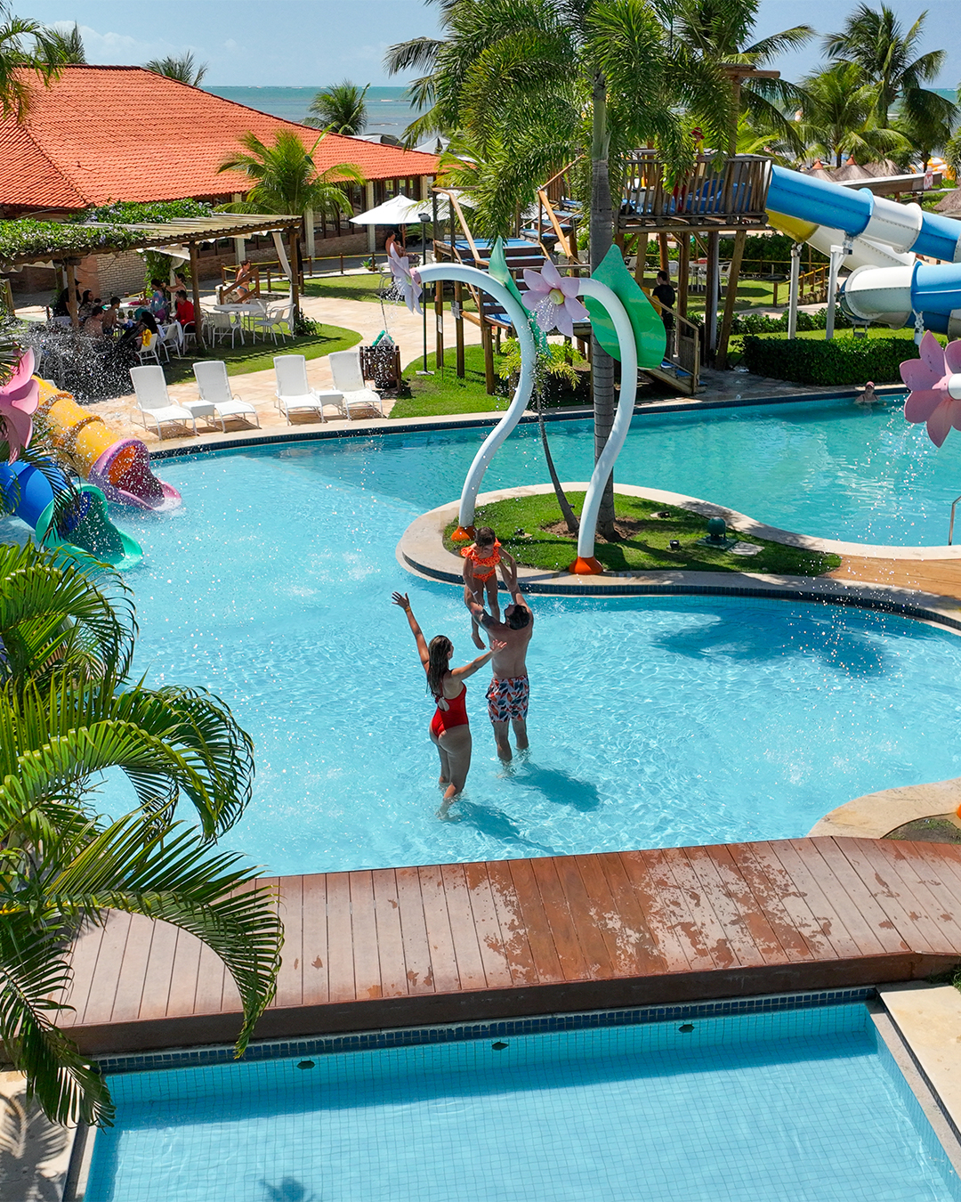 Família na piscina infantil onde há um pequena ponte de madeira do Salinas Maragogi. O pai segura a filha nos braços e a mãe está de braços abertos dentro da piscina para crianças do resort.