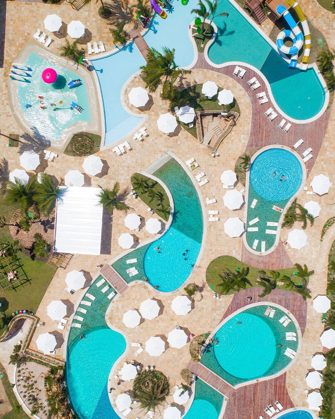 Área externa do Salinas Maragogi mostrando as principais piscinas, as infantis com os brinquedos. Com várias espreguiçadeiras, guarda-sóis. A estrutura do resort é cercada por coqueiros e gramas verdes.