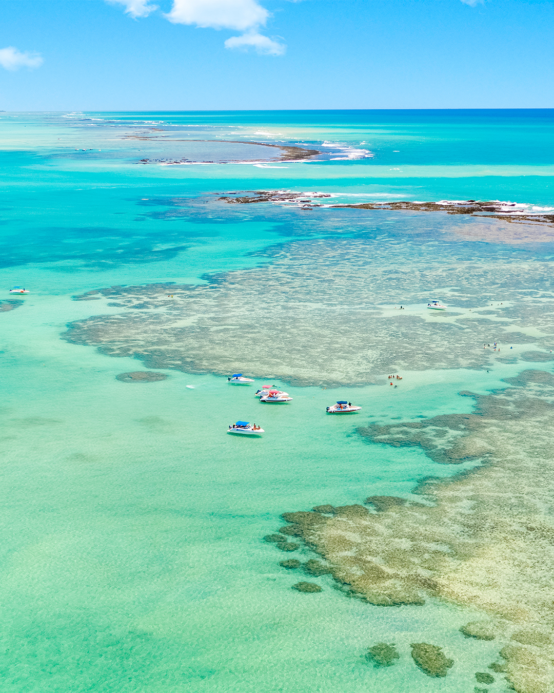Vista aérea de Barra Grande, as famosas piscinas naturais, com seus recifes de corais formando águas rasas e cristalinas de cor turquesa em alto mar, com vários barcos e turistas no local sob um céu azul.