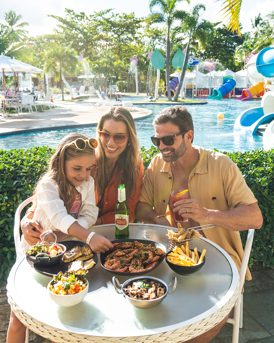 Família sorrindo e almoçando à beira da piscina em um dia ensolarado. A mesa redonda está cheia de pratos com camarões, batatas fritas e saladas. O pai segura um drink e a mãe está com a filha no colo e tem uma garrafa de cerveja na mesa. Ao fundo, coqueiros, a piscina e um playground aquático colorido.