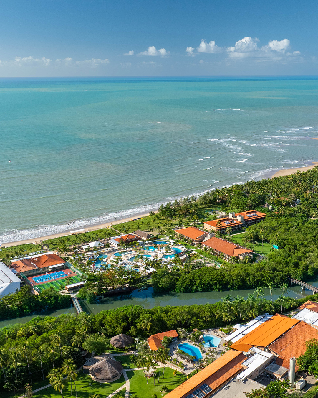 Vista de ângulo elevado do complexo do Salinas Maragogi, mostrando uma área de lazer infantil, quadras, piscinas maiores, estrutura do resort, com o rio cortando o resort com uma ponte ligando os dois lados, e o mar azul com a praia ao fundo, tudo cercado por coqueiros.