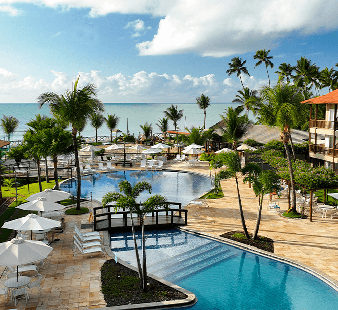 Resort à beira-mar com edifícios de telhados marrons. Há múltiplas piscinas, espreguiçadeiras e guarda-sóis brancos. Palmeiras altas e o oceano são visíveis ao fundo.