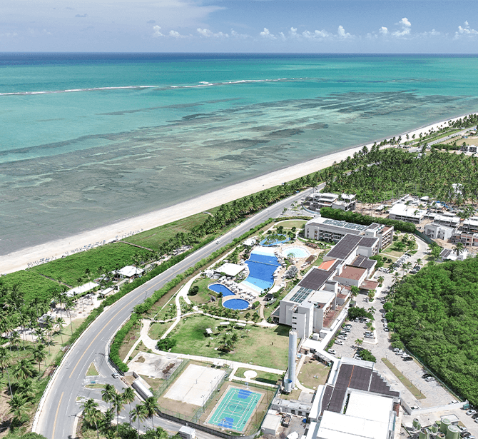 Vista aérea do Japaratinga Resort, ao redor está o oceano, faixa de areia branca com guarda-sóis, piscina em forma de lagoa e edifícios modernos, coqueiros e vegetação também, criando um cenário tropical.