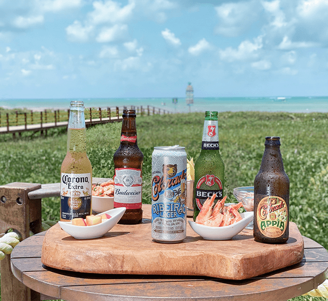 Uma mesa de madeira à beira-mar apresenta várias bebidas: Corona, Budweiser, Becks, Colorado Appia e Cerveja Beira. Ao lado, um prato com camarões e frutas. Ao fundo, uma vista do oceano e um farol distante. A atmosfera é relaxante e convidativa.