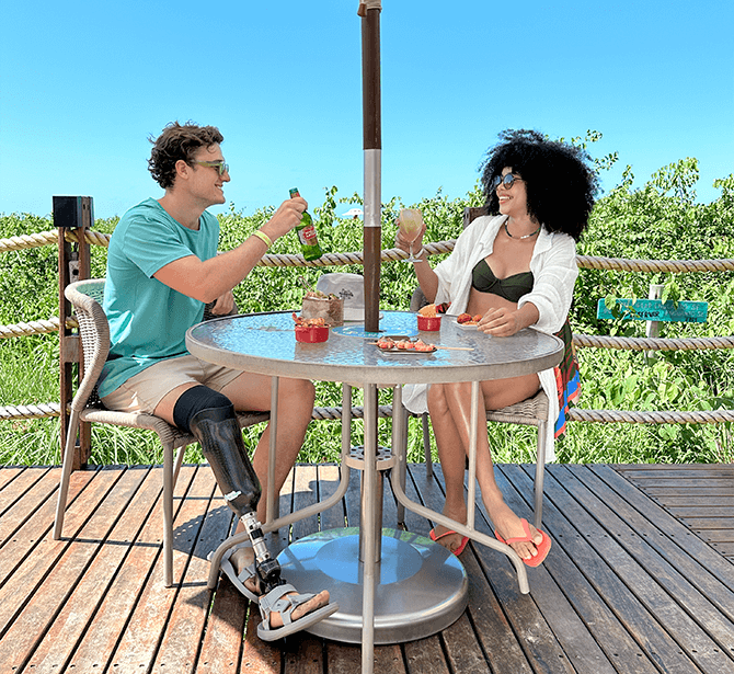 Esta é uma imagem de duas pessoas brindando ao ar livre. O homem usa óculos e tem uma prótese na perna. A mulher tem cabelo afro volumoso. Ambos estão em um ambiente relaxante, possivelmente com vista para a natureza.